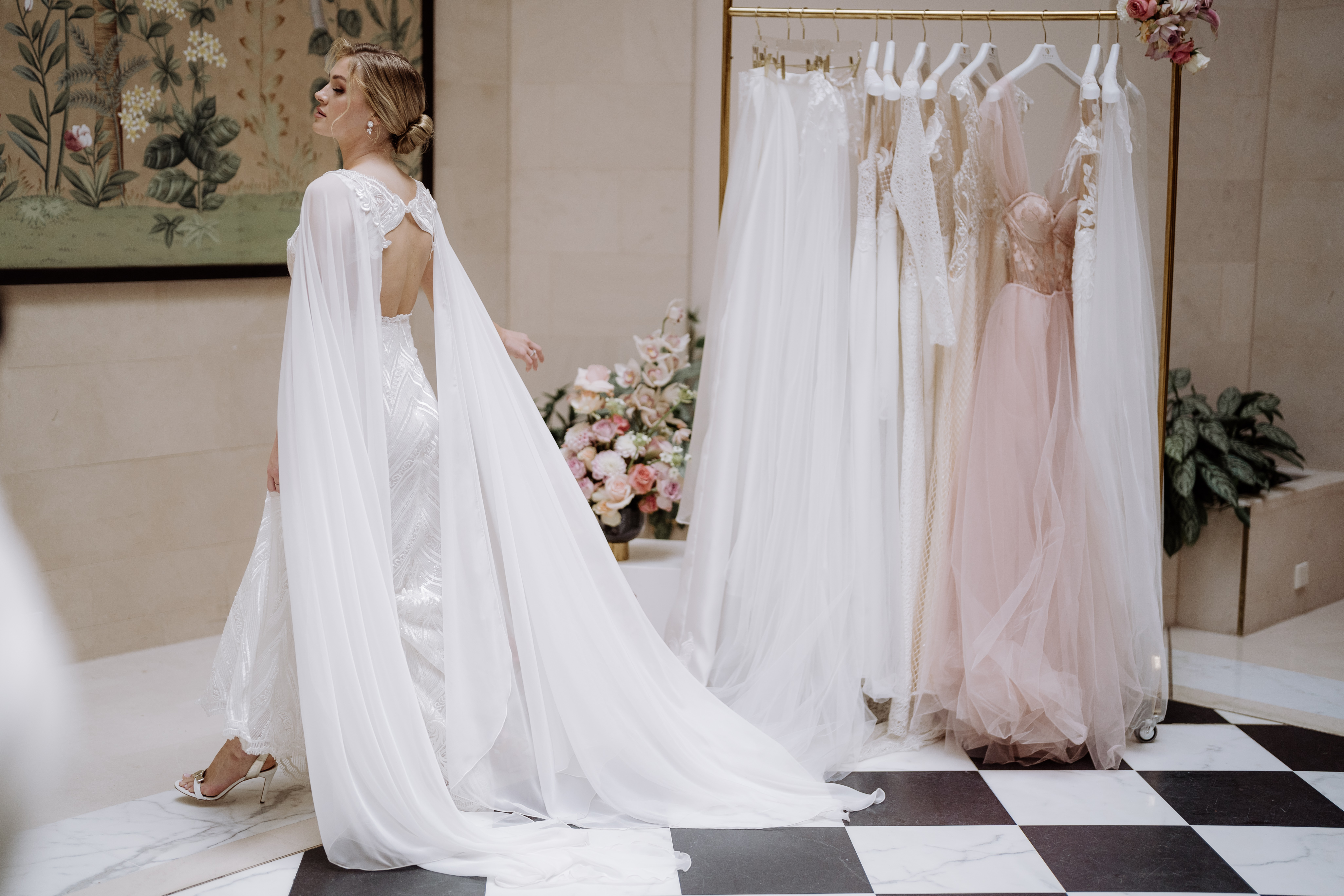 Wie finde ich mein perfektes Brautkleid? 10 goldene Regeln, die alles perfekt machen. 1 Braut mit weißem Cape auf schwarz-weiß gefliestem Boden neben einem Kleiderständer mit Brautkleidern und einem rosefabenen Kleid.