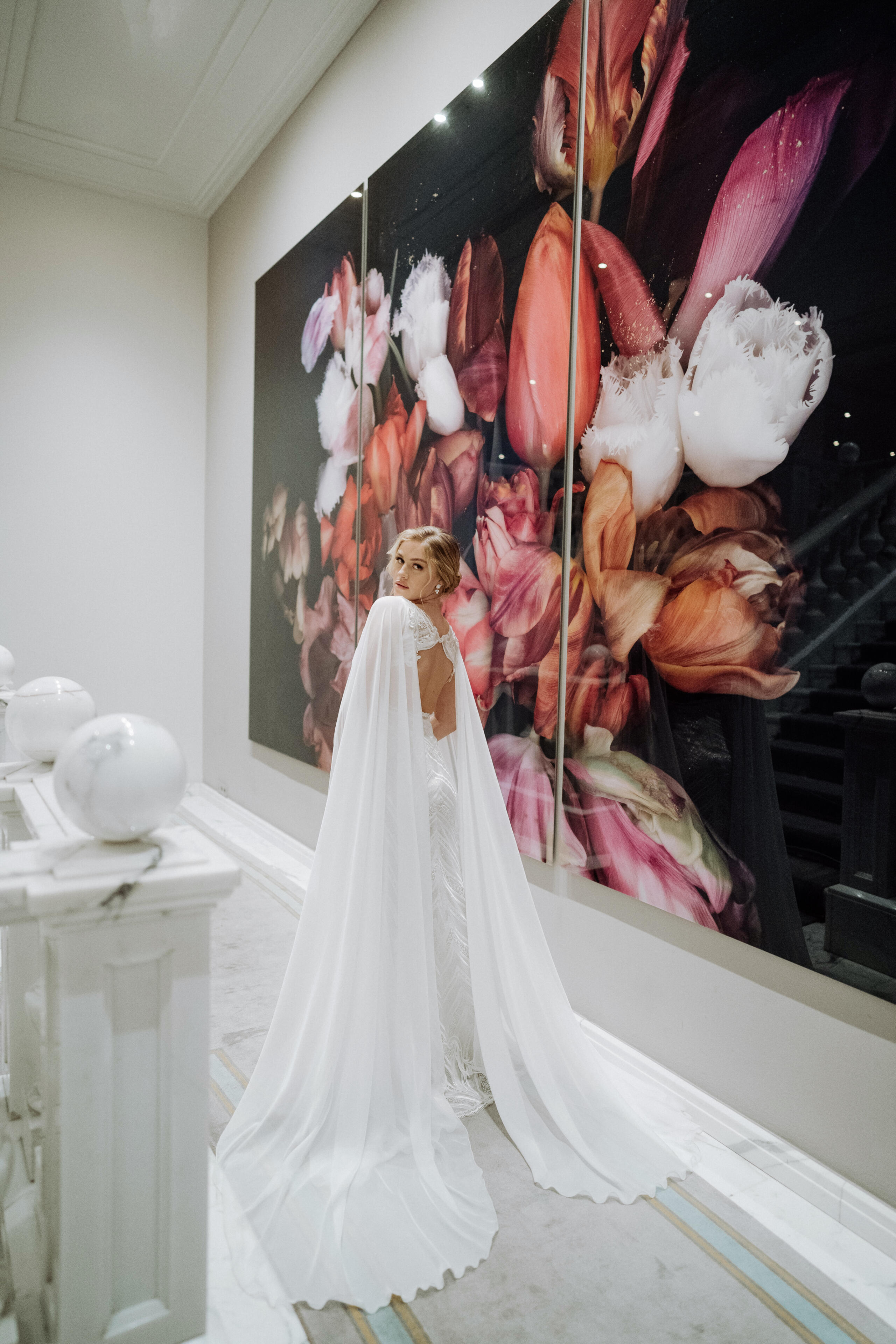 Wie finde ich mein perfektes Brautkleid? 10 goldene Regeln, die alles perfekt machen. 3 Blonde Braut mit luftigem Brautcape, die sich nach Hinten dreht, steht vor einem großen Bild mit rosafarbenen Tulpen.