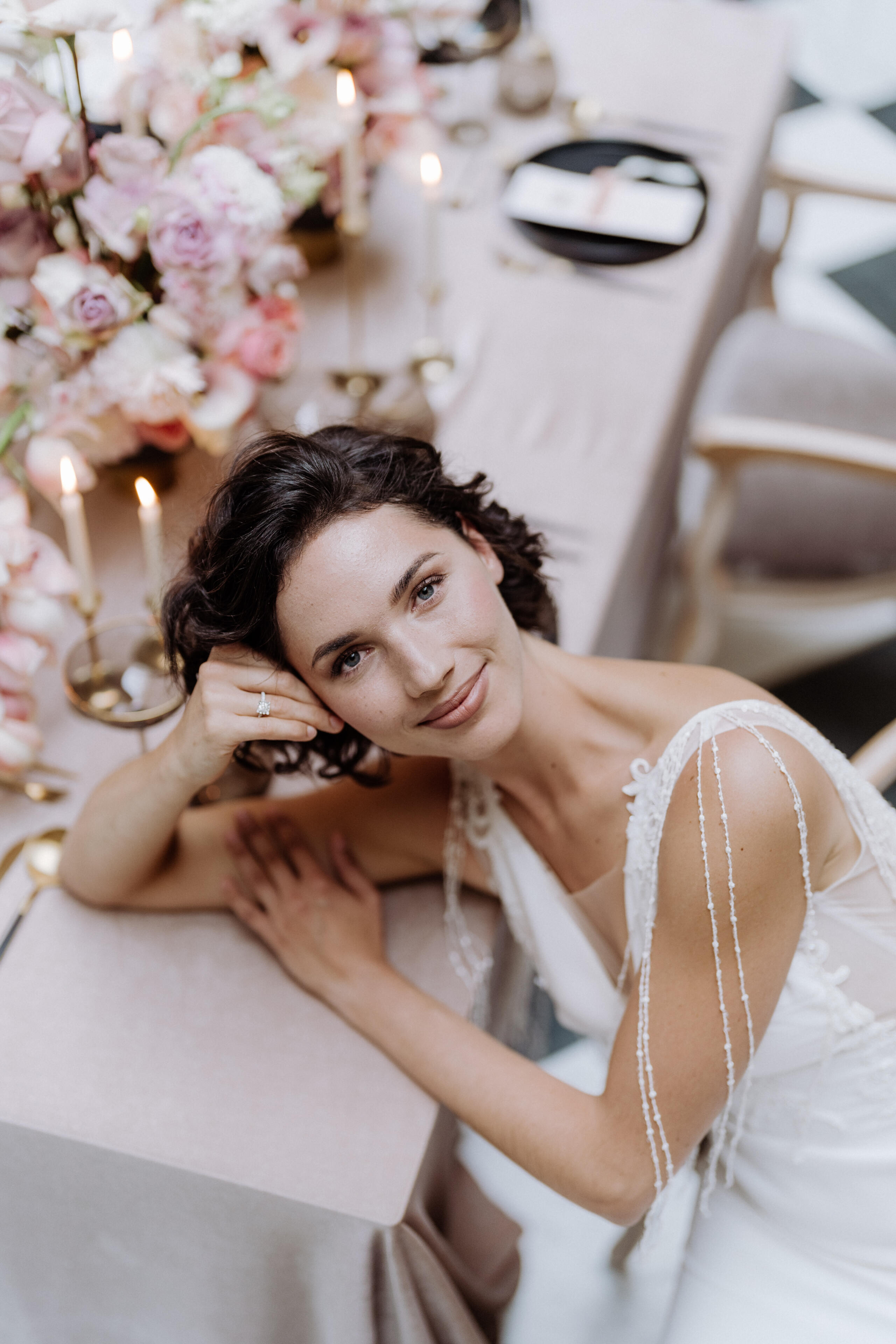 Wie finde ich mein perfektes Brautkleid? 10 goldene Regeln, die alles perfekt machen. 5 Braut mit schwarzen, leicht lockigen Haaren sitzt im Brautkleid am Tisch, stützt ihr Gesicht auf die Hand und blickt verträumt nach oben.